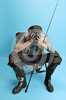 Portrait of young man, fisherman with fishing rod, spinning and equipment sitting isolated over blue studio background
