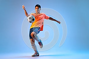 Portrait of young man, football player in motion, action, kicking ball with knee isolated over blue studio background in