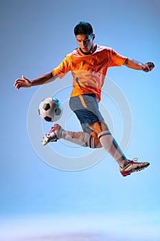 Portrait of young man, football player in motion, training, kicking ball in a jump isolated over blue studio background