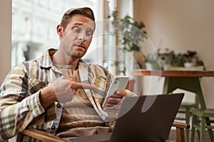 Portrait of young man freelancer, working from cafe, sitting on chair with laptop, pointing finger at his phone screen