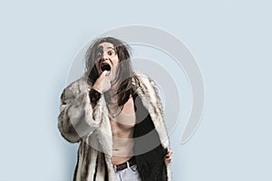 Portrait of young man in fur coat with finger in mouth standing against light blue background