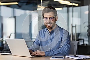 Portrait of a young man in glasses sitting at a desk in the office and working on a laptop, looking and smiling at the