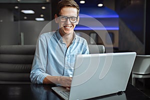 Portrait of a young man with a good mood, a businessman in a shirt and glasses, who works on a laptop in a cafe, can be used for a