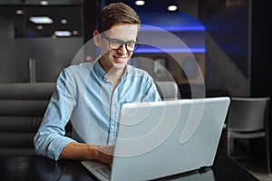 Portrait of a young man with a good mood, a businessman in a shirt and glasses, who works on a laptop in a cafe, can be used for a