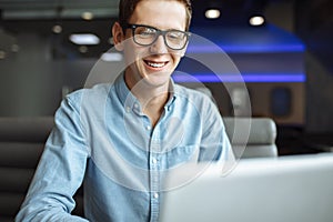 Portrait of a young man with a good mood, a businessman in a shirt and glasses, who works on a laptop in a cafe, can be used for a