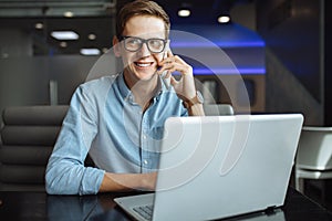 Portrait of a young man with a good mood, a businessman in a shirt and glasses, who works on a laptop and talking on the phone in