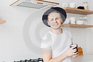 Portrait young man in hat smiles drink fresh coffee in ceramic black mug breakfast. Bright kitchen background