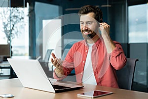 Portrait of a young man in a headset. He sits in the office, works on a laptop, video call