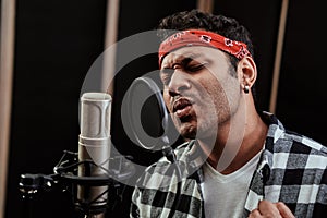 Portrait of young man, hip hop artist looking focused, singing into a condenser microphone while recording a song in a