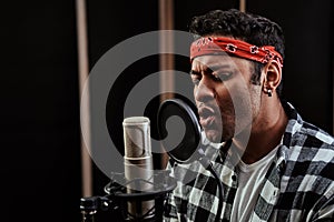 Portrait of young man, hip hop artist looking focused, singing into a condenser microphone while recording a song in a