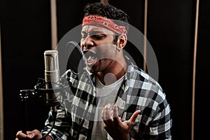 Portrait of young man, hip hop artist singing with closed eyes into a condenser microphone while recording a song in a