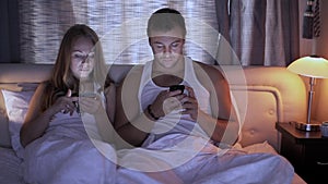 Portrait of a young man and his wife both using their smartphones while lying together in bed