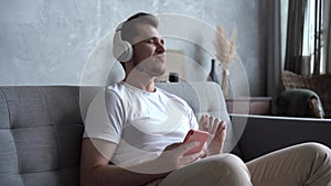 Portrait of young man listening to music in headphones using smart phone sitting on sofa at home.