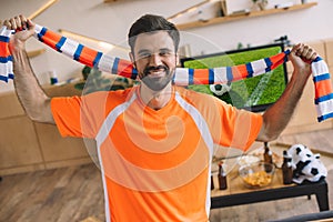 portrait of young man in orange fan t-shirt holding striped scarf and celebrating victory in soccer match
