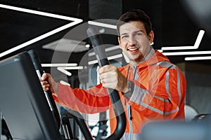 Portrait of a young man in orange windbreaker workout on a fitness machine at gym.