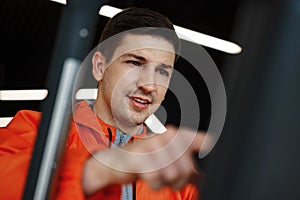 Portrait of a young man in orange windbreaker workout on a fitness machine at gym.