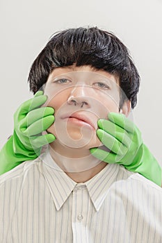 Portrait of young man with piercing posing with hands in rubber gloves on face  over grey studio background