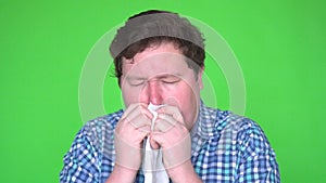 Portrait of young man in plaid shirt with allergy or cold, blowing his nose with a tissue, isolated green screen.