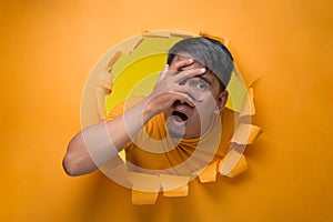 Portrait of young man poses through torn paper hole, covering his face by hand and looking at camera