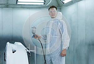 Portrait young man of professional car painter with protective clothing standing next to car in auto paint room and with gun