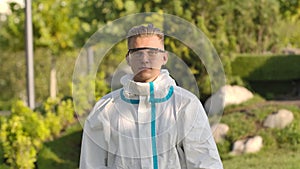 Portrait of a young man in a protective suit, glasses and gloves looks at the camera and holds a smartphone with green