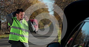 Portrait of young man in a safety vest standing in front of his broken car, looking at car engine and talking on cell