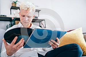 Portrait of young man sitting in comfortable sofa in living room taking out 12-inch vinyl LP from sleeve. Old vintage analog sound