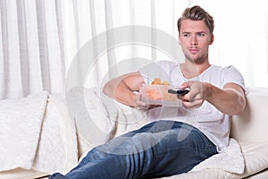 Portrait young man sitting on couch and eating chips and zapping
