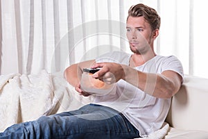 Portrait young man sitting on couch and eating chips and zapping