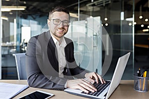 Portrait of a young man sitting at a desk in an office center in a suit and working on a laptop. Looking and smiling at