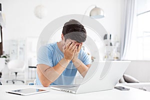 Portrait of young man sitting at his desk at home with a laptop sad covering his face with his hands. Freelance and selfemployment photo