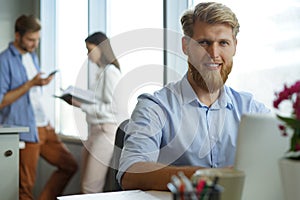 Portrait of young man sitting at his desk in the office, working on his laptop.