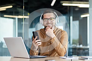 Portrait of a young man sitting in the office at the desk wearing headphones, working on a laptop and holding a phone