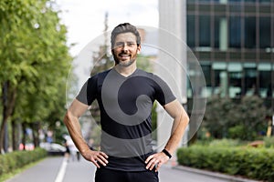 Portrait of a young man in sportswear standing outside on the street, hands on waist, confidently and smilingly looking photo