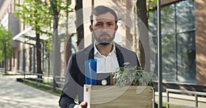 Portrait of young man in suit holding box with his stuff while standing at street. Male office worker in 30s lost his