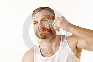 Portrait of young man in T-shirt, taking care of his facial skin using massage scrapper against white background