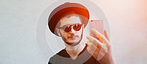 Portrait of young man taking selfie, standing over white background. Dressed in black, wearing sunglasses and hat.