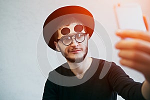 Portrait of young man taking selfie, standing over white background. Dressed in black, wearing sunglasses and hat.