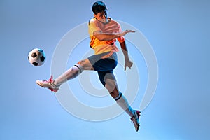 Portrait of young man in uniform, professional football player kicking ball in a jump isolated over blue studio