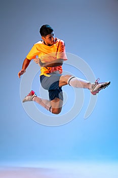 Portrait of young man in uniform, professional football player kicking ball in a jump isolated over blue studio