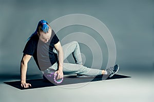 Portrait of a young man with an unusual hairstyle of mohawk from blue braids in sportswear. Doing leg exercise by dint of roller f