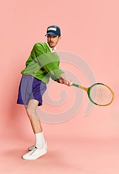 Portrait of young man in vintage sportive clothes posing with tennis racket isolated over pink background. Active