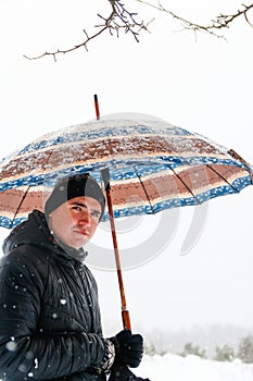 Portrait of young man in warm hat outside on rural winter snowy background holding umbrella. Happy millennial serious