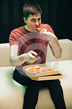 Portrait of young man watching TV eating pizza sitting on sofa at home. Relaxation cinema channels resting. No emotions