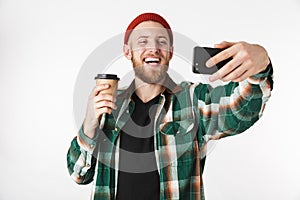 Portrait of young man wearing plaid shirt holding paper cup with coffee and using mobile phone, while standing isolated over white