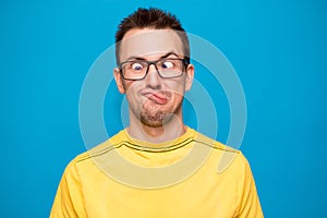Portrait of young man wearing yellow shirt and trendy nerd glasses isolated over blue with confused face and funny expression
