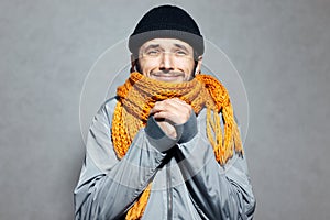 Portrait of young man who feels cold, wearing orange scarf and black beanie hat, on gray background.