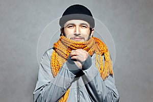 Portrait of young man who feels cold, wearing orange scarf and black beanie hat, on gray background.