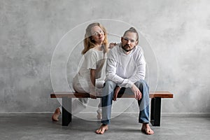 Portrait of a young man and woman in white clothes and without shoes, sitting on a bench on a minimalist background.