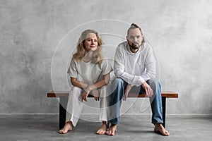 Portrait of a young man and woman in white clothes and without shoes, sitting on a bench on a minimalist background.